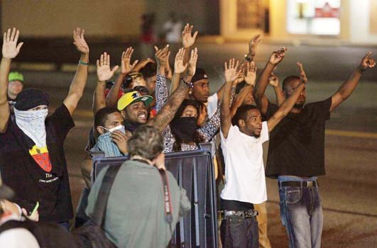 Protesters of the shooting death of Michael Brown Jr. take the “hands up, don’t shoot” stance in Ferguson, Mo. as police point their weapons at them.