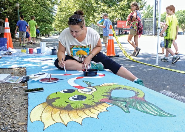 Abby Klein creates a work of art Friday atop a storm drain on Tredegar Street near Brown’s Island as curious passers-by watch her progress. She is one of five artists selected by the Richmond Department of Public Utilities from 55 for the first phase of the Storm Drain Art Project. Ms. Klein’s work is titled, “You drop it, I drink it.” The other four artists who are painting drains along Tredegar Street are Alfonso Perez Acosta, Nicole Gomez, Melissa Greedy and Richard Lucente. 
The city is paying a commission of $300 to each artist and later plans to award a $200 prize to the artist whose work the public votes as their favorite. The project was set up as a fun, creative approach to educate the public to keep the drains free of litter and not use the drains to get rid of oil and other toxins that could pollute the city’s waterways and, ultimately, the James River into which storm water flows. 