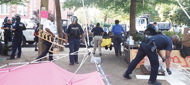 Richmond Police officers remove belongings on July 30 from “Grace Park,” a two-month-old encampment created by protesters, after residents of the area complained. Location: The grassy median at Allen Avenue
and Grace Street north of the Lee statue on Monument Avenue. On the same day, police also cleared a similar camp on the Monument Avenue median north of the camp. Two people were arrested. By Monday, activists were once again distributing food, water and literature from tables in the Allen Avenue medians. Participants said police officers were regularly checking, but mostly appeared to condone the activity. However, they said police issued warnings against overnight stays on the medians. Police reported that about 11:15 p.m. July 30, shots were fired into a vehicle near the Lee statue circle following an altercation and that the glass
door of a residence in the 1600 block of Monument Avenue had been shattered by reported gunfire. Officers canvassing the area found a rifle and ammunition magazines. “Gunfire and violent behavior is not peaceful, nonviolent protest; it is criminal, unacceptable and will not be tolerated in our city,” Richmond Police Chief Gerald M. Smith said in a statement. “The City of Richmond will take all necessary steps to protect residents and visitors and preserve peace and public safety in our communities.”