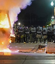 Police stand near a garbage truck ablaze during protests, Monday, Aug. 24, 2020, in Kenosha, Wis., sparked by the shooting of Jacob Blake by a Kenosha Police officer a day earlier. Officials in Kenosha, Wisconsin, enforced their curfew selectively, targeting demonstrators protesting police brutality while allowing "militia members" and supporters of law enforcement to roam the street, a federal lawsuit alleges./Credit:	Morry Gash/AP