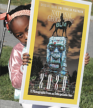 Storie Nzassi, the 7-year-old granddaughter of Delegate Delores L. McQuinn of Richmond, holds
an enlarged copy of the cover of National Geographic Magazine’s January edition featuring a projection of George Floyd’s face
on the statue of Confederate Gen. Robert E. Lee in Richmond. The Monument Avenue statue became a rallying point for protesters against police brutality and racial injustice following Mr. Floyd’s death in May
at the hands of a Minneapolis police officer. Images representing the struggle for civil rights and equality in the United States were projected onto the monument by Richmond artists Dustin Klein and Alex Criqui and photographed by Kris Graves for the cover. The youngster was attending a news conference last Friday with her grandmother at the Virginia Museum of Fine Arts, where Gov. Ralph S. Northam proposed $11 million in state funds be used to transform Monument Avenue.