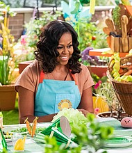 On a rooftop garden, Michelle Obama, wearing a blue apron, converses with a furry white and blue puppet with frozen waffle ears, a round, pink mochi puppet, and a bee puppet wearing a red tie around a table dressed with green decorations for a birthday party.
Credit: Adam Rose/Netflix