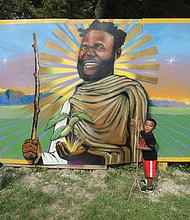 “That’s my daddy,” said 5-year-old Anais Lewis of Roanoke, while staring at the mural of his father, John R. Lewis, whose image and life was memorialized on May 26 at the Sankofa Community Orchard, 301 Covington Road on Richmond’s South Side. Mr. Lewis, who died in January 2021, taught students how to grow their own food and spent time “hanging out” in the garden, often keeping youngsters out of harm’s way as they got their hands dirty together. Mr. Lewis was the co-founder and executive director of Renew Richmond, a nonprofit that operated several urban agricultural sites and sometimes had pop-up stands where fresh produce that was grown and harvested by local Black youthS and adults was also often sold by them too. He advocated for Black communities to understand the power of gardening and self-sufficiency.
Duron Chavis, the founder of the Sankofa Community Garden, said Mr. Lewis was one of the “founding fathers of Black urban agriculture in the City of Richmond.” The mural was created by lead painter Sir James L. Thornhill assisted by R. Vashti Woods, both of Richmond.
