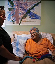 Constance Guthrie, who is 74, sits in a hospital-style bed propped up by a pillow. Her daughter, Jessica Guthrie, stands next to the bed, smiling. She was diagnosed with Alzheimer’s disease at age 66. About 14% of Black people in America over the age of 65 have Alzheimer’s, compared with 10% of white people, according to the Centers for Disease Control and Prevention. The disparity is likely even more, because many Black people aren’t correctly diagnosed.