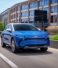 Front 7/8 view of a 2024 Chevrolet Equinox EV LT in Riptide Blue Metallic with a Summit White roof driving in a downtown area.