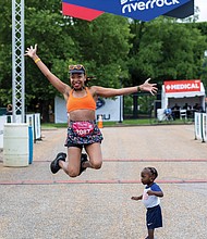April Burwell jumps for joy in front of the finish line after completing the Bust the Banks Half Marathon trail run at Dominion Energy Riverrock on Sunday, May 18. Her 19-month-old son, Julian, stands grounded beside her, taking it all in.