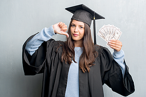 Young brunette woman over grunge grey wall wearing graduate uniform holding dollars with angry face, negative sign showing dislike with thumbs down, rejection concept