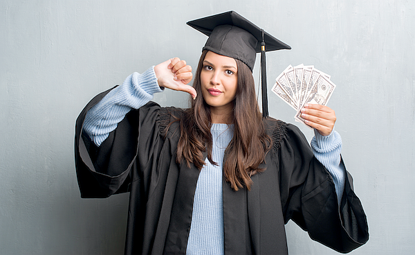 Young brunette woman over grunge grey wall wearing graduate uniform holding dollars with angry face, negative sign showing dislike with thumbs down, rejection concept
